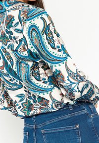 Vit blus med mönster av paisleyblommor i blått, orange och svart, matchad med blå denimjeans, visad från bak- och sidovy.