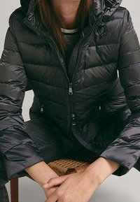Giacca a vento nera con cappuccio, con design trapuntato, chiusura frontale con zip e tasche laterali. Il tessuto appare liscio e lucido.