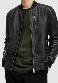 Svart läderbomberjacka med dragkedja framtill, ribbade manschetter och krage. Har en bröstficka och slät textur, bärs över en grön stickad topp.