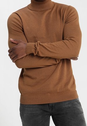 Osoba ubrana w brązowy sweter z golfem, ze skrzyżowanymi ramionami, w ciemnoszarych dżinsach na jednolitym białym tle.