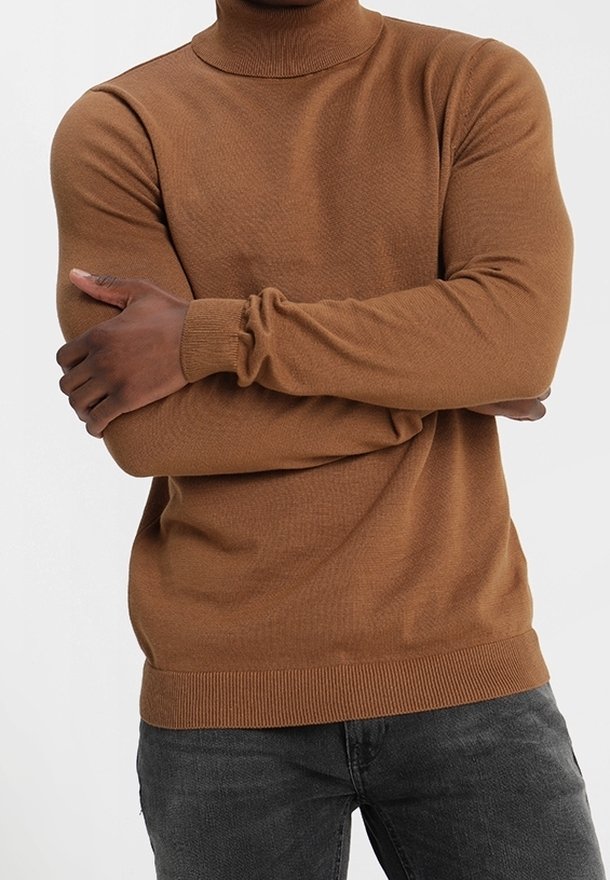 Personne portant un pull à col roulé marron, les bras croisés, associée à un jean gris foncé sur un fond blanc uni.