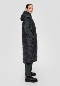 Schwarze Steppweste mit Kapuze, mit einem gesteppten Muster, seitlichen Taschen und langem Schnitt. Getragen über einem grünen Pullover und dunklen Hosen.