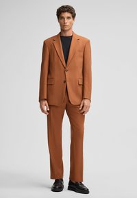 Oranje textuurpak met een enkelvoudig gebreide blazer en recht моделiger broek. Gedragen over een zwart shirt, gecombineerd met zwarte schoenen.
