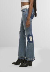 Femme portant un jean flare taille haute avec des patchworks et des bords effilochés, associé à des bottines à talons noires et un top court blanc.