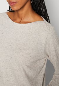 Femme aux cheveux tressés portant un haut à manches longues gris clair moucheté avec un large col rond, sur un fond neutre.