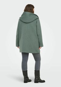 Manteau en laine mélangée vert avec un grand col châle, des manches longues et une coupe décontractée, associé à un jean skinny gris foncé et des bottes noires.