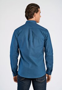 Pitkähihainen sininen paita, jossa napillinen kaulus; takana on olkasauma ja laskosyksityiskohta, valmistettu teksturoidusta kankaasta.