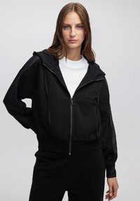 Sudadera con cremallera negra de acabado texturizado, con capucha, dos bolsillos frontales y mangas largas con paneles transparentes. Llevada sobre una camiseta blanca.