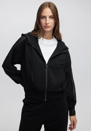Sudadera con cremallera negra de acabado texturizado, con capucha, dos bolsillos frontales y mangas largas con paneles transparentes. Llevada sobre una camiseta blanca.