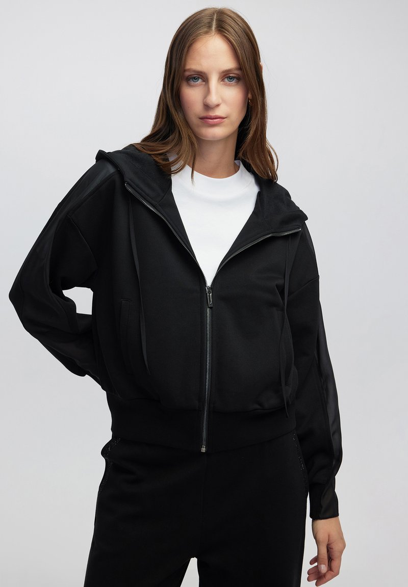 Sudadera con cremallera negra de acabado texturizado, con capucha, dos bolsillos frontales y mangas largas con paneles transparentes. Llevada sobre una camiseta blanca.