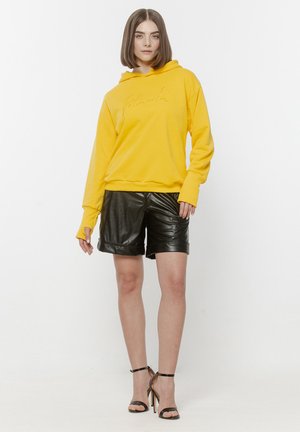 Kobieta ubrana w żółtą bluzę z kapturem, czarne skórzane krótkie spodenki i czarne sandały na wysokim obcasie z paskiem na kostce, stojąca na białym tle.