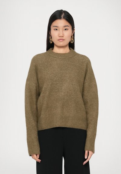 Jeune femme avec des cheveux noirs raides portant des boucles d'oreilles en chaîne dorée, un pull en laine bouclée vert olive et un pantalon noir, se tenant contre un fond blanc.