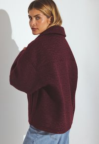 Burgundy oversized jacket with a wide collar, textured fabric, drop shoulders, and side pockets, paired with light blue denim jeans.