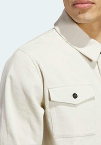 Chaqueta de pana beige claro con cuello doblado, cierre de cremallera y un bolsillo en el pecho con detalle de botón negro.