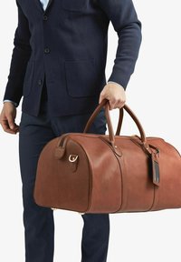Brun läder resväska med rundad form, dubbla handtag, guldfärgad hårdvara och en rektangulär etikett fäst. Texturerad yta.