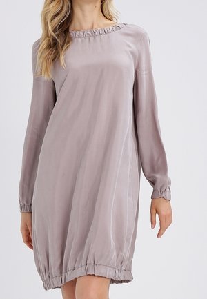 Femme portant une robe ample à manches longues de couleur mauve clair, avec des poignets et un ourlet élastiqués, sur un fond uni.