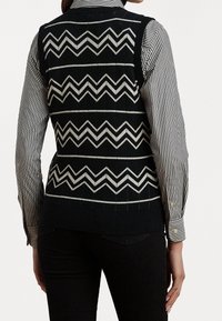 Czarna dziergana kamizelka z kontrastującym, białym wzorem zygzaka. Noszona na czarno-białej koszuli w paski z mankietami na guziki.