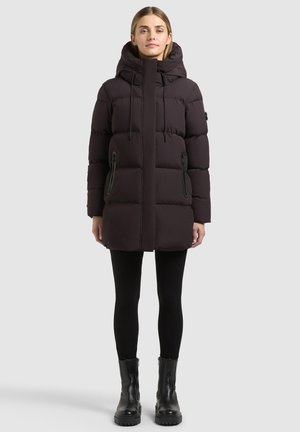 Femme portant un long manteau noir matelassé avec capuche, des leggings noirs et des bottines noires debout devant un fond uni.