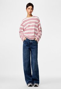 Woman wearing a white long-sleeve shirt with red stripes, wide-leg blue jeans, and sneakers, standing with hands in pockets against white background.