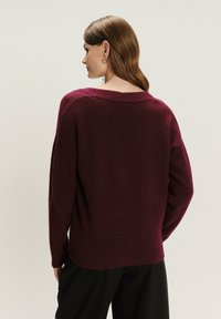 Vrouw met steil bruin haar, draagt gouden hoepelringen en een losse donkerpaarse trui, gezien van achteren tegen een lichte achtergrond.