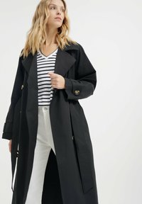 Długi czarny płaszcz trench z szerokim kołnierzem, złotymi guzikami i wcięciem w talii. Nosi się go na paskowanym, v-neck t-shircie i białych spodniach.