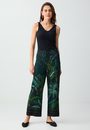 Femme debout portant un haut noir sans manches, un pantalon vert ample avec de grands motifs de feuilles tropicales, et des chaussures noires faciles à enfiler.