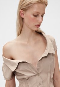 Beige skjorta med knappar framtill, korta ärmar som är avsmalnande vid axlarna och en djup urringning. Mjukt tyg med slät textur och diskret glans.