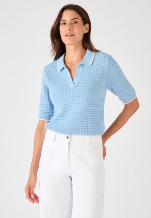 Frau trägt ein strukturiertes, hellblaues, kurzärmeliges Strick-Poloshirt und weiße, hochtaillierte Hosen, steht mit einer Hand in der Hüfte.