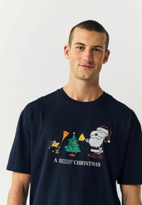 Tumedat sinise puuvillane T-särk, millel on värvikas graafika Snoopy ja Jõuluvana, puu ning muusikariistadega. Tekst ütleb "Snoopy Jõulud."