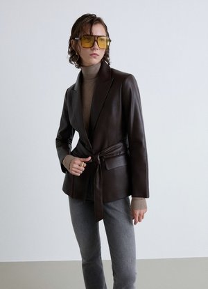 Donna con blazer in pelle nera con cintura, dolcevita beige, pantaloni grigi e grandi occhiali da sole con lenti gialle, in piedi davanti a uno sfondo semplice.