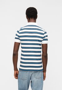 Άνδρας που φοράει λευκό πόλο μπλουζάκι με μαύρες και μπλε οριζόντιες ρίγες και ανοιχτόχρωμο μπλε τζιν, στέκεται γυρισμένος με την πλάτη σε απλό φόντο.