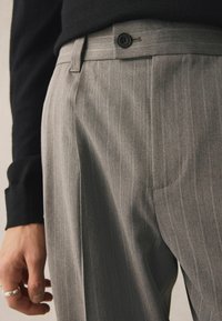 Pantalons en chevrons gris avec une texture lisse, dotés d'une fermeture à bouton unique et de poches latérales, portés avec un pull noir.