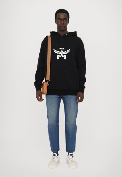 Haut à capuche noir avec logo MCM blanc et motif de laurier, assorti à un jean bleu et des baskets blanches. Le modèle porte également un sac bandoulière marron.