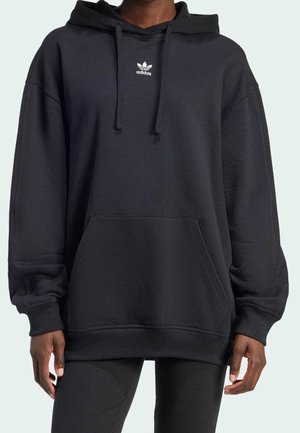 Person iført en sort Adidas hættetrøje med snøre, en front-kængurulomme og et lille hvidt logo på brystet.