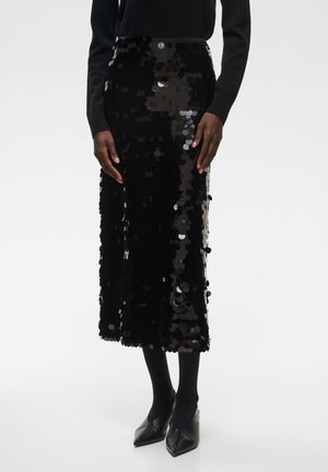 Personne portant un haut noir à manches longues, une jupe midi noire à sequins, des collants noirs et des chaussures noires à bout pointu, debout devant un fond uni.
