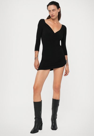 Černé obepnuté šaty s dlouhými rukávy, výstřihem do V, přiléhavým střihem a textilní strečovou tkaninou. Nosené s černými koženými kozačkami po kolena.
