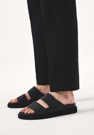 Lábak fekete papucsot viselnek két széles pánttal és fekete nadrággal, világos háttér előtt.