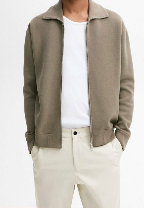 Bruine jas met rits en kraag, gemaakt van zacht stof. Gedragen over een wit T-shirt, gecombineerd met beige broek. Subtiele stikdetails.