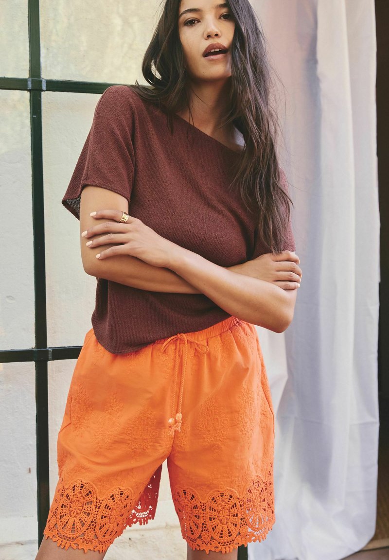 Frau mit langen dunklen Haaren trägt ein braunes kurzärmeliges Oberteil und orange bestickte Shorts, steht mit verschränkten Armen drinnen.