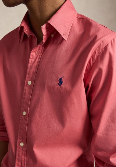 Άνδρας που φοράει κοραλί πουκάμισο με κουμπιά και μικρό σκούρο μπλε κεντημένο λογότυπο παίκτη πόλο στο στήθος.