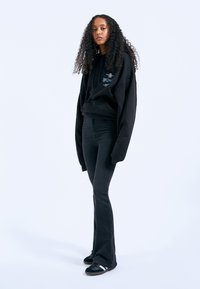Black oversized hoodie with embroidered design, paired with fitted black flared jeans and black sneakers with white stripes.