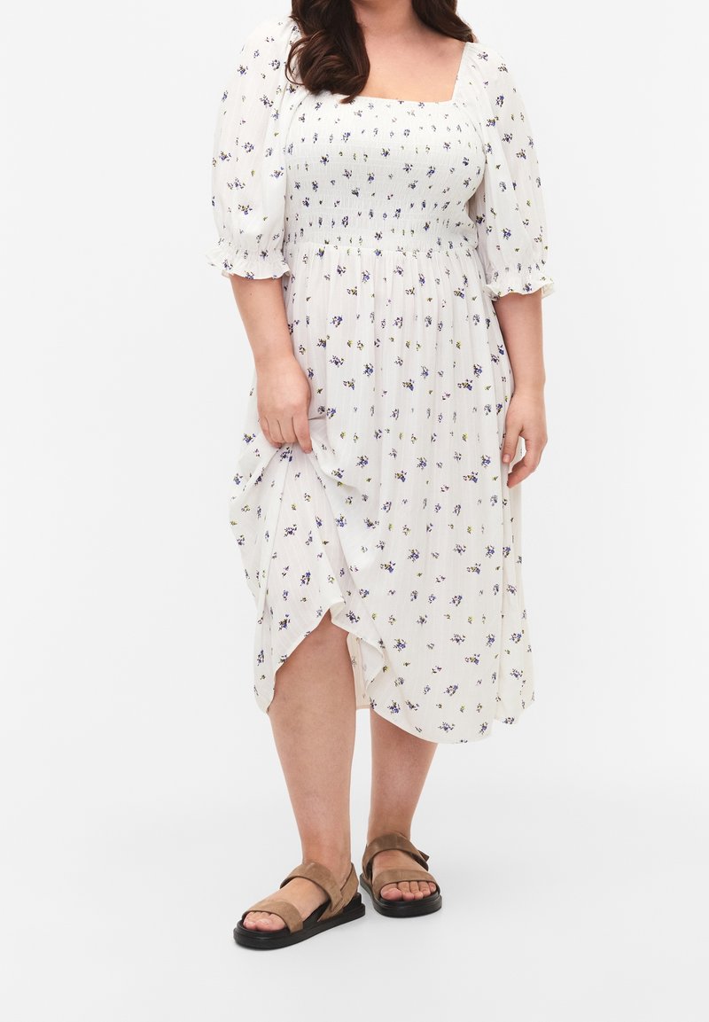 Bílé květinové šaty se čtvercovým výstřihem, krátkými puff rukávy, elastickým nařasením v pase a bočním rozparkem; doplněné o hnědé sandály.