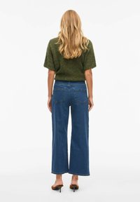 Top de punto verde combinado con vaqueros azul de pierna ancha. El suéter tiene mangas cortas, mientras que los pantalones cuentan con un diseño clásico de cinco bolsillos.