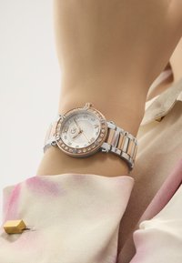Reloj metálico en plata y oro rosa con una esfera redonda, adornado con cristales y números romanos, en una muñeca junto a una suave tela de pastel.