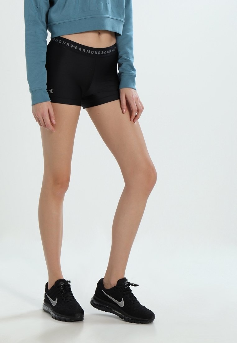 Donna che indossa pantaloni corto neri Under Armour, un maglione corto blu e scarpe da corsa nere Nike, in piedi davanti a uno sfondo bianco.