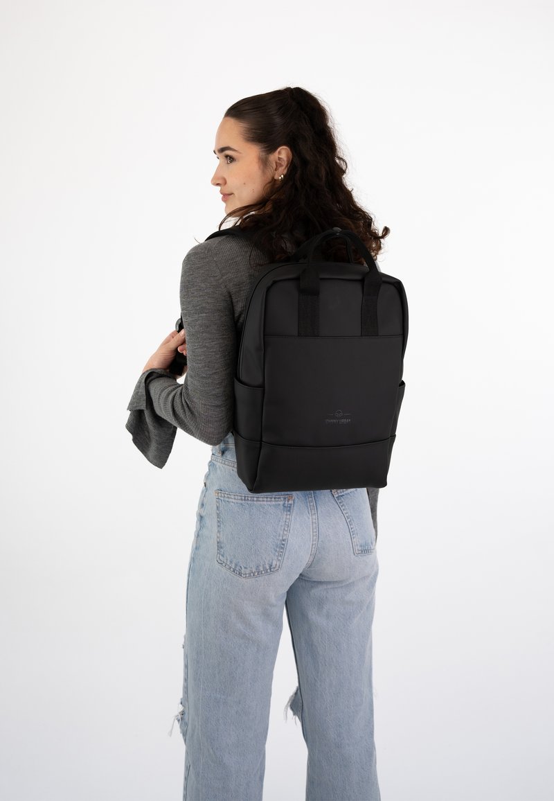 Mochila negra con correas de hombro acolchadas, textura suave y un diseño sencillo. Cuenta con bolsillos frontales y un detalle de logo en la parte posterior.