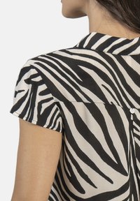 Camisa de manga corta con estampado de cebra en blanco y negro, con un cuello tipo solapa y un detalle de botón en la parte posterior. Textura de tela suave.