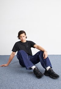 Svart t-shirt med liten blå logotyp, lösa blå denimbyxor och svarta sneakers med vita strumpor. Modell sitter på blå matta.