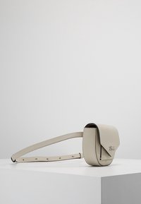 Vaaleanbeige nahkainen olkalaukku, jossa on jämäkkä muoto, magneettinen läpällinen suljin ja säädettävä hihna, jossa on pieniä metallisia yksityiskohtia.