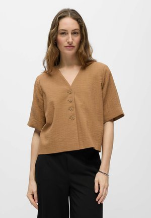 Frau mit schulterlangem gewelltem Haar, die ein beigefarbenes, geknöpftes V-Ausschnitt-Kurzarm-Top und schwarze Hose vor weißem Hintergrund trägt.